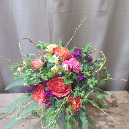 Colorful arrangement of orange and pink roses with purple flowers and curly branches in a low vase