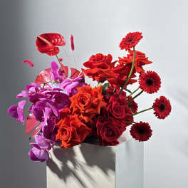 Red and purple floral arrangement with roses and gerbera daisies in a white pedestal vase