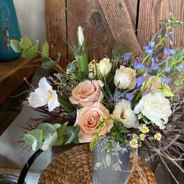 Peach roses, white blooms, and purple delphinium in a ribbed gray vase on a woven mat.