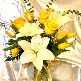 Yellow roses and white lilies arranged in a glass vase with a gold ribbon.