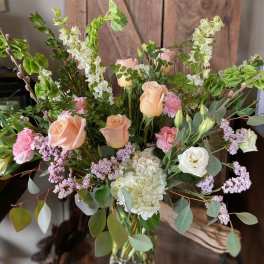 Tall arrangement of peach roses, white hydrangea, pink flowers and green bells of Ireland in a glass vase