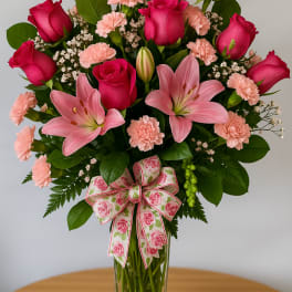 Tall arrangement of hot pink roses, pink lilies, and light pink carnations in a clear vase with a floral bow.