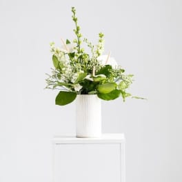 White and green floral arrangement in a tall white ribbed vase on a white pedestal