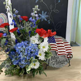 Patriotic floral arrangement with blue hydrangeas, red carnations, and white daisies beside a flag plaque