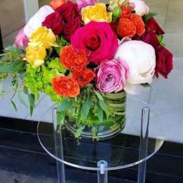 Low glass vase of bright roses, peonies, and spray roses in pink, red, orange, and yellow on a clear stand.