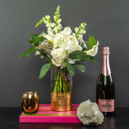 White floral arrangement in a glass vase with a champagne bottle and candle holder