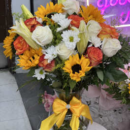 Bouquet of sunflowers, roses, lilies, and white daisies in a glass vase with a yellow bow