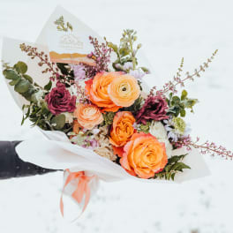 Hand-tied bouquet of orange and yellow blooms with burgundy accents wrapped in white paper.