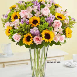 Bouquet of pink roses, sunflowers, and purple flowers in a clear glass vase