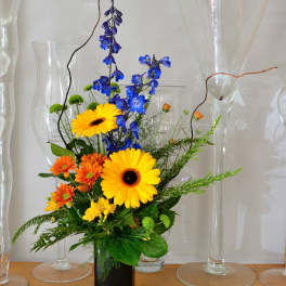 Tall arrangement of yellow gerbera, orange mums, and blue delphinium in a black rectangular vase