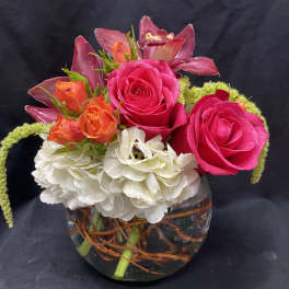 Short arrangement of hot pink roses, orange spray roses, orchids and white hydrangea in a glass bowl vase
