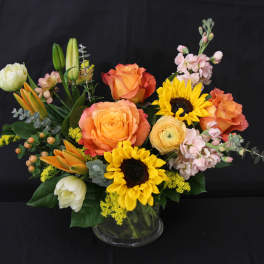 Mixed bouquet with orange roses, sunflowers, and pink blooms in a glass vase