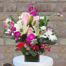 Compact arrangement of pink roses, white lilies and bright pink blooms in a green glass cube vase