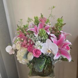 Tall arrangement of pink lilies, white orchids, roses, and hydrangeas in a clear glass vase