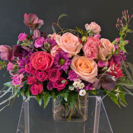 Low rectangular bouquet of pink, peach, and purple flowers in a clear glass vase