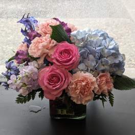 Low arrangement of pink roses, peach carnations and blue hydrangeas in a clear glass cube vase