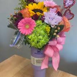 Colorful bouquet with a sunflower, gerbera daisies, and a pink ribbon in a purple vase