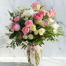 Pink and white bouquet of roses, carnations, and daisies in a glass vase with a ribbon.