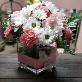 Pink carnations and white daisies in a clear square vase with a ribbon