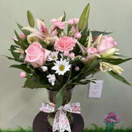 Pink roses, lilies, carnations, and white daisies in a clear glass vase with a floral bow.