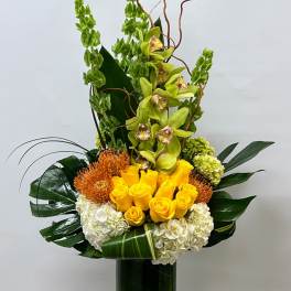 Tall floral arrangement with yellow roses, green orchids, and white hydrangeas in a glass vase