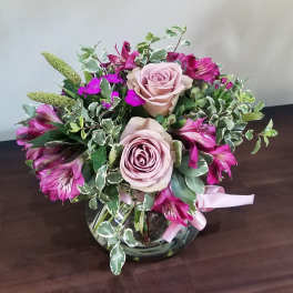 Round bouquet of mauve roses and magenta alstroemeria in a clear glass bowl vase with pink ribbon