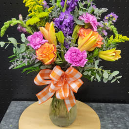 Mixed bouquet of orange roses, yellow lilies, and purple flowers in a glass vase