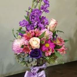 Tall mixed bouquet of purple and pink flowers with roses and daisies in a glass vase tied with a lavender bow