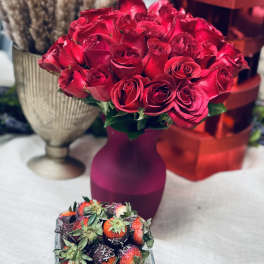 Red rose bouquet in a purple vase with chocolate-covered strawberries in front