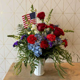 Patriotic arrangement with red roses, red carnations, blue hydrangeas and a small American flag in a white vase