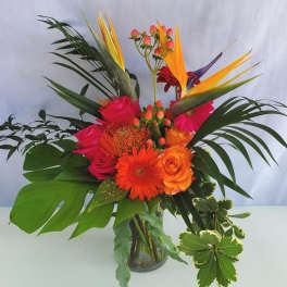 Tall tropical arrangement with orange gerberas, roses, and birds of paradise in a clear glass vase.