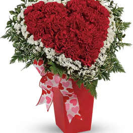 Heart-shaped red carnation arrangement in a red vase with a heart ribbon