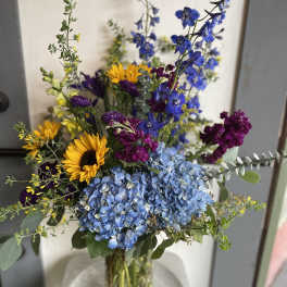 Mixed bouquet with blue hydrangeas, sunflowers, and purple blooms in a glass vase