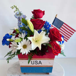 Red roses and white lilies in a patriotic wooden container with a small U.S. flag.