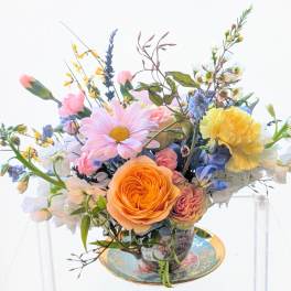 Pastel arrangement of roses, daisies, and mixed blooms in a patterned teacup on a matching saucer