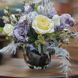 Short arrangement of cream roses and purple ruffled flowers in a dark glass vase