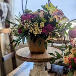 Basket-style arrangement of purple roses and white blooms with green accents in a brown container.