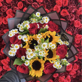 Handheld bouquet of red roses, yellow sunflowers, and white daisies wrapped in black paper over a bed of red roses.