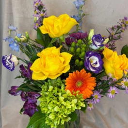 Mixed bouquet with yellow roses, orange gerbera daisy, and purple blooms in a glass vase
