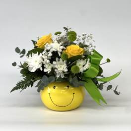 Yellow and white flower arrangement in a smiley-face pot with a green ribbon