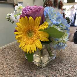 Colorful bouquet with a yellow gerbera daisy, purple rose, and blue hydrangea in a glass vase