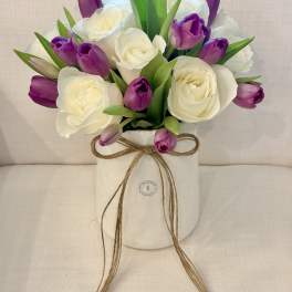 White roses and purple tulips arranged in a white vase with a twine bow.