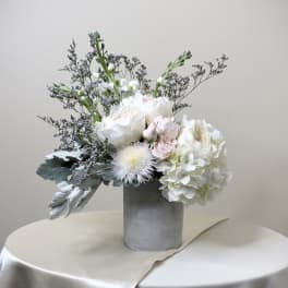 Asymmetrical arrangement of white and blush flowers in a gray cylinder vase on a draped ivory table.