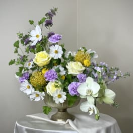 Garden-style arrangement of white, yellow, and lavender flowers in a metal pedestal bowl on a draped table.