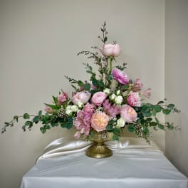 Asymmetrical arrangement of pink and peach roses and ranunculus in a gold pedestal vase with trailing greenery.