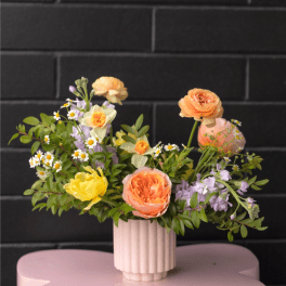 Low pastel arrangement of peach and yellow flowers in a ribbed vase on a pink table