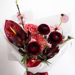 Hand-tied bouquet of dark red ranunculus, pink roses, and tropical blooms wrapped in white paper with burgundy ribbon