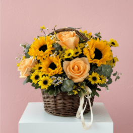 Basket arrangement of yellow sunflowers, peach roses, and small yellow daisies with a ribbon bow.
