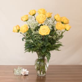 Tall arrangement of yellow roses and white filler flowers in a clear glass vase on a wooden surface