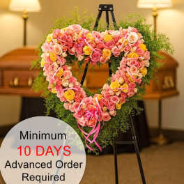 Heart-shaped wreath of pink and yellow roses on a metal easel in a funeral setting.